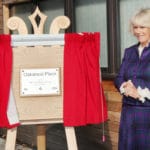 Camilla, Duchess of Cornwall standing next to a wooden easel with red curtains and a plaque reading 'Oakwood Place'.