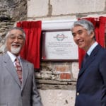 Two men in suits smiling beside a Honda Racing 50th Anniversary plaque mounted on a stone wall with red curtains.